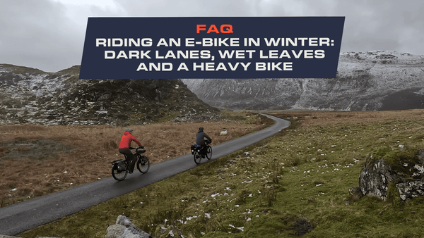 Two riders on loaded e-bikes on a wet single-track lane below snow-covered mountains in Snowdonia